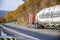 Brown classic bonnet big rig semi truck with bulk semi trailer go downhill on the winding autumn road with yellow maples on the