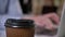 Brown cup of coffee on table in office, hands of businessman are typing on laptop on background, blurred background
