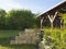 brown tiber wooden gazebo or pergola with climbing plants and white flowers, surrounding sandstone wall, green grass and bush in