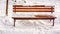 Brown wooden bench with knots with white fresh snow on it in winter park with a lantern on a background