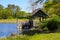 A brown wooden boat house on a gorgeous vast blue lakes surrounded by lush green trees, grass and plants with blue sky