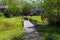 A brown wooden bridge over a silky green lake surrounded by lush green trees, grass and plants