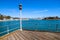 A brown wooden pier with a gray metal hand railing and a tall grey and red lamp post surrounded by vast deep blue ocean water