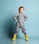 Brunet child in orange hat, gray overall, yellow rubber boots. He is smiling, hands on hips, posing on blue studio background
