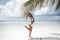 Brunette Bikini Model jumping by palm on sand on tropical beach vacation. Happy traveller woman. Sexy slim girl in white swimwear