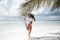 Brunette Bikini Model jumping by palm on sand on tropical beach vacation. Happy traveller woman. Sexy slim girl in white swimwear