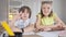 Brunette Caucasian boy raising hand and talking as girl sitting at desk looking at camera. Children studying online on