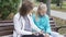 Brunette caucasian grandmother and her young blond granddaughter sitting at the bench and talking. Teenage girl