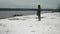 Brunette in dark green coat and pink sport shoes stands on snowy bank of frozen river.