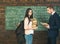 Brunette female student standing in front of green board with heap of books and huge backpack. Professor looking at