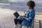 Brunette girl with afro hair works with her laptop sitting on a park bench on a spring day