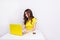 A brunette girl with curly hair looks frustrated sitting in front of laptop in yellow case. She sits behind white table