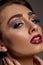 Brunette girl in earrings posing sideways on gray background. Colorful eyeshadow, false eyelashes, glossy purple lips