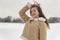 Brunette girl poses beautifully in fashionable beige sheepskin coat decorated with white fur on snowy glade background