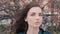 Brunette girl posing with blank epression in front of cherry tree in bloom and wind play with her long hair