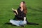 Brunette girl sits cross-legged on a green lawn and laughs joyfully while reading.