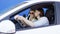 Brunette man and blonde woman are taking photos inside white car against blue background. Funny driver and his cheerful