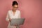 A brunette stands on a pink background with collected hair in glasses and looks into a laptop while holding it in front