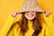 A brunette stands on a yellow background in a yellow sweater, pulling a hat over a half-shirt and smiles