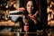 Brunette woman barman carefully pours cocktail from steel shaker into glass using sieve.