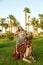 A brunette woman in a cape on her head firmly clings to the lying camel on the grass against the backdrop of palm trees.