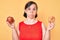 Brunette woman with down syndrome holding red apple and donut sitting puffing cheeks with funny face