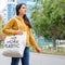 A brunette woman in an orange hoodie walks with a fabric bag that says No more plastic in the city