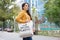 A brunette woman in an orange hoodie walks with a fabric bag that says No more plastic in the city