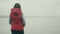 Brunette woman in red jacket stands over misty river in drizzly rain, view back