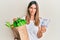 Brunette young woman holding groceries and 50 colombian pesos banknotes relaxed with serious expression on face