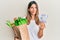 Brunette young woman holding groceries and 50 colombian pesos banknotes smiling looking to the side and staring away thinking