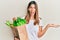 Brunette young woman holding paper bag with bread and groceries celebrating achievement with happy smile and winner expression