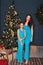 Brunettes, mom and daughter in identical blue dresses are standing near the New Year tree indoor in the christmas decorated room