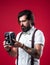 Brutal bearded man in suspenders holding retro camera while standing against red background, photography