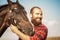 Brutal guy with a beard leads a horse. Farmer in a plaid shirt and