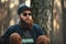 A brutal young man with a huge beard in sunglasses and a cap in the woods at sunset.