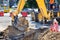 The bucket of a large construction excavator digs a trench on the road against the backdrop of a city street on a sunny summer day