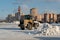 A bucket loader removes snow from snowdrifts in a city square after a snowfall against the backdrop of residential buildings on a