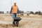 Builder in an orange reflective vest leading a wheelbarrow throw road construction field