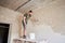 Builder is standing on wooden stand and working with spatula with plaster at the wall in small room