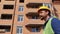 A builder in a yellow helmet with a beard and mustache goes near the house under construction and tells about the construction pro