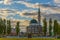 Building of mosque with minaret stands between pyramidal poplars against background of blue summer sky in rays of sunset.