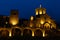 The building of an old stone monastery that looks like a castle. At night, the building is illuminated in bright yellowish colors