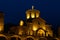 The building of an old stone monastery that looks like a castle. At night, the building is illuminated in bright yellowish colors