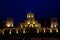 The building of an old stone monastery that looks like a castle. At night, the building is illuminated in bright yellowish colors