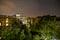 Buildings and green trees at night, illuminated cloudy sky