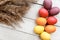 A bunch of colored eggs are laying on a white wooden table beside some wheat spikelets closeup