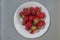 A bunch of red strawberries lying on top of each other in a white ceramic plate that stands on a grayish-silver wicker tablecloth