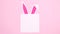 Bunny ears appear under paper card note on pastel pink background. Easter stop motion flat lay