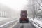 Burgundy big rig semi truck tractor without semi trailer driving on the street winter road with fog and snow